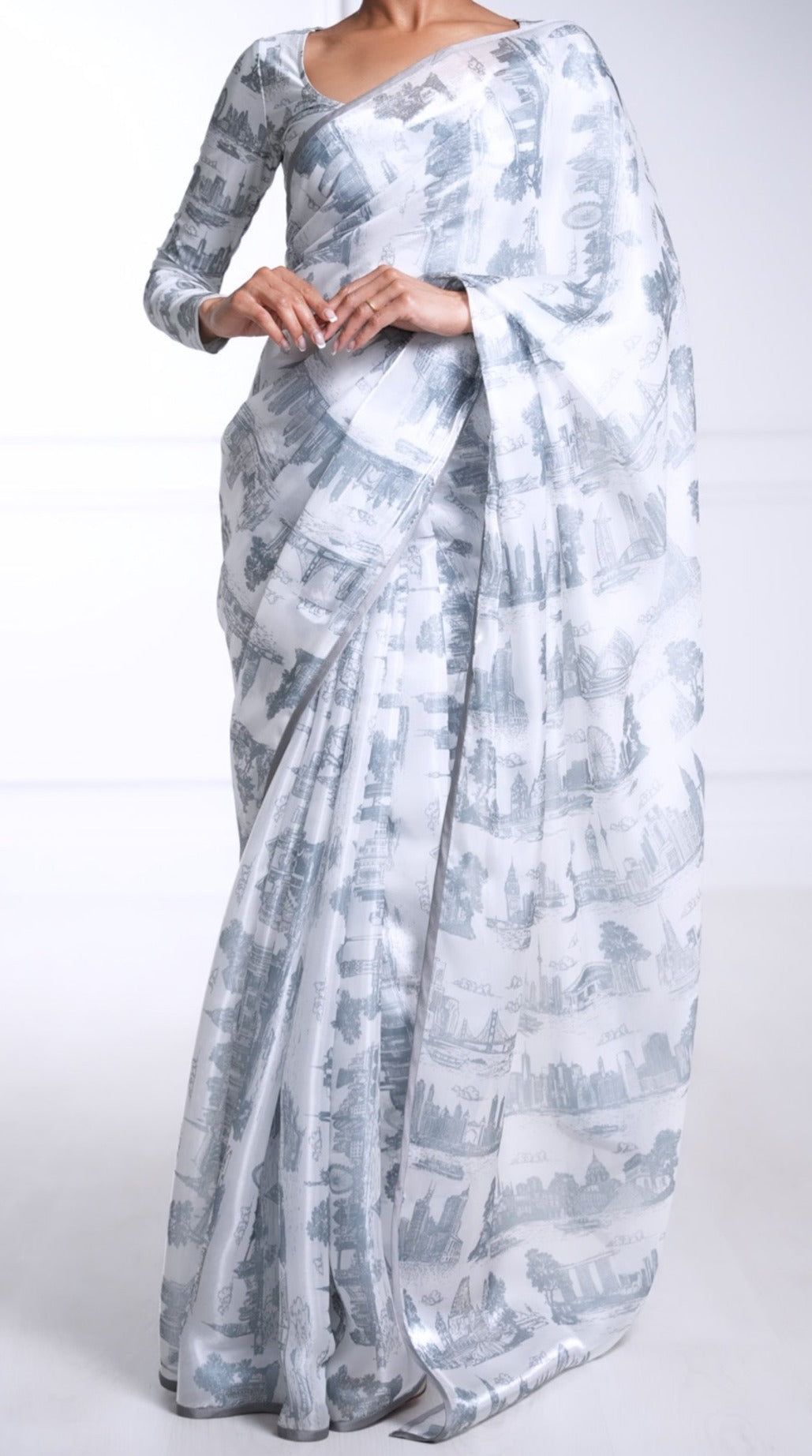 Model draped in a white organza saree with a grey satin border and grey printed city skylines throughout. Model is also wearing a matching printed long sleeve blouse with sweetheart neckline.