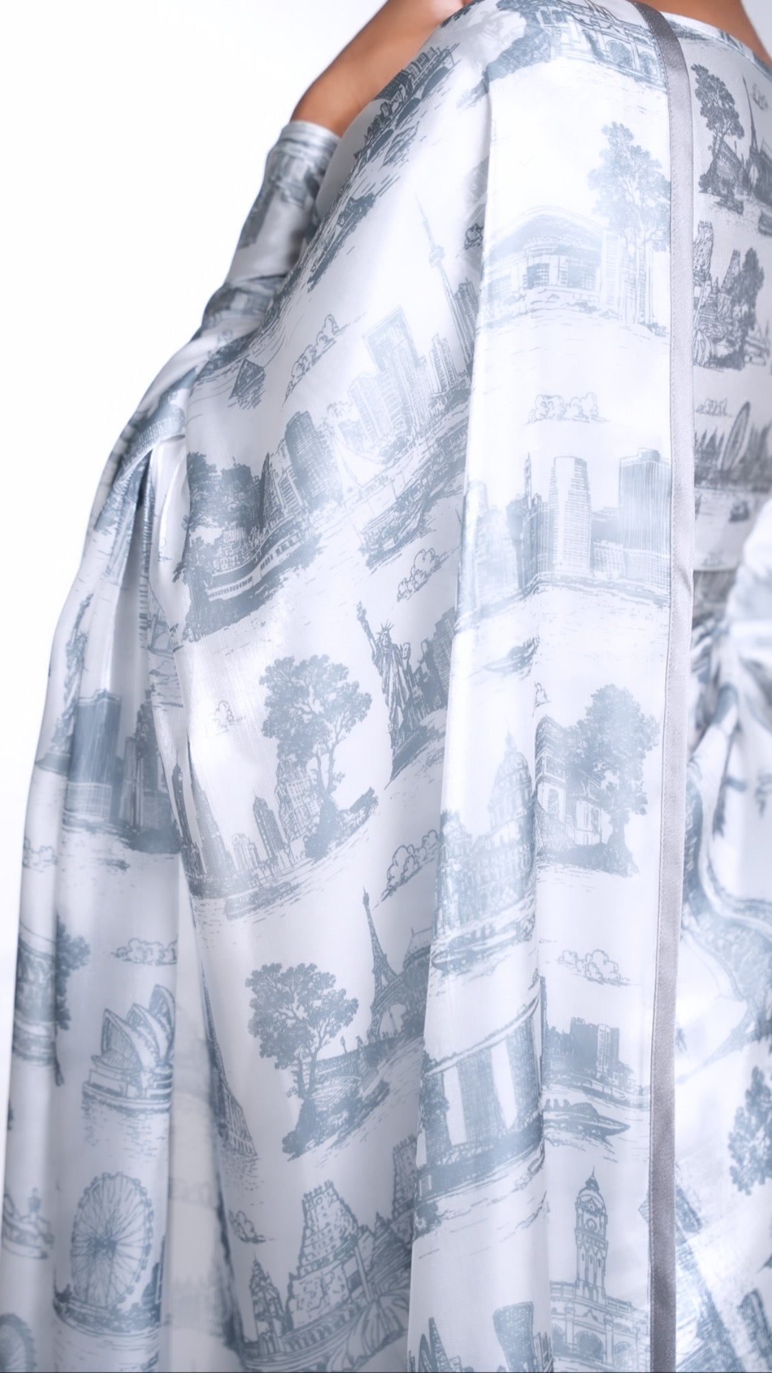 Close side view of a model draped in a white organza saree with a grey satin border and grey printed city skylines throughout. Model is also wearing a matching printed long sleeve blouse.