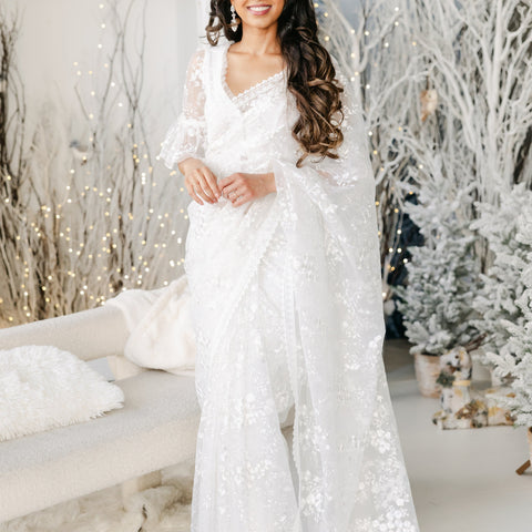 Model draped in a white transparent mesh saree with white floral embroidery and a white lace border. Model is wearing a matching white blouse with flared sleeves and a white saree petticoat underneath. Christmas themed background.