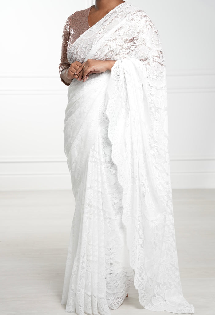 Model draped in a white lace saree with scalloped edging. Model is also wearing a 3/4 sleeve v-neck rose gold crop top and white saree petticoat underneath.