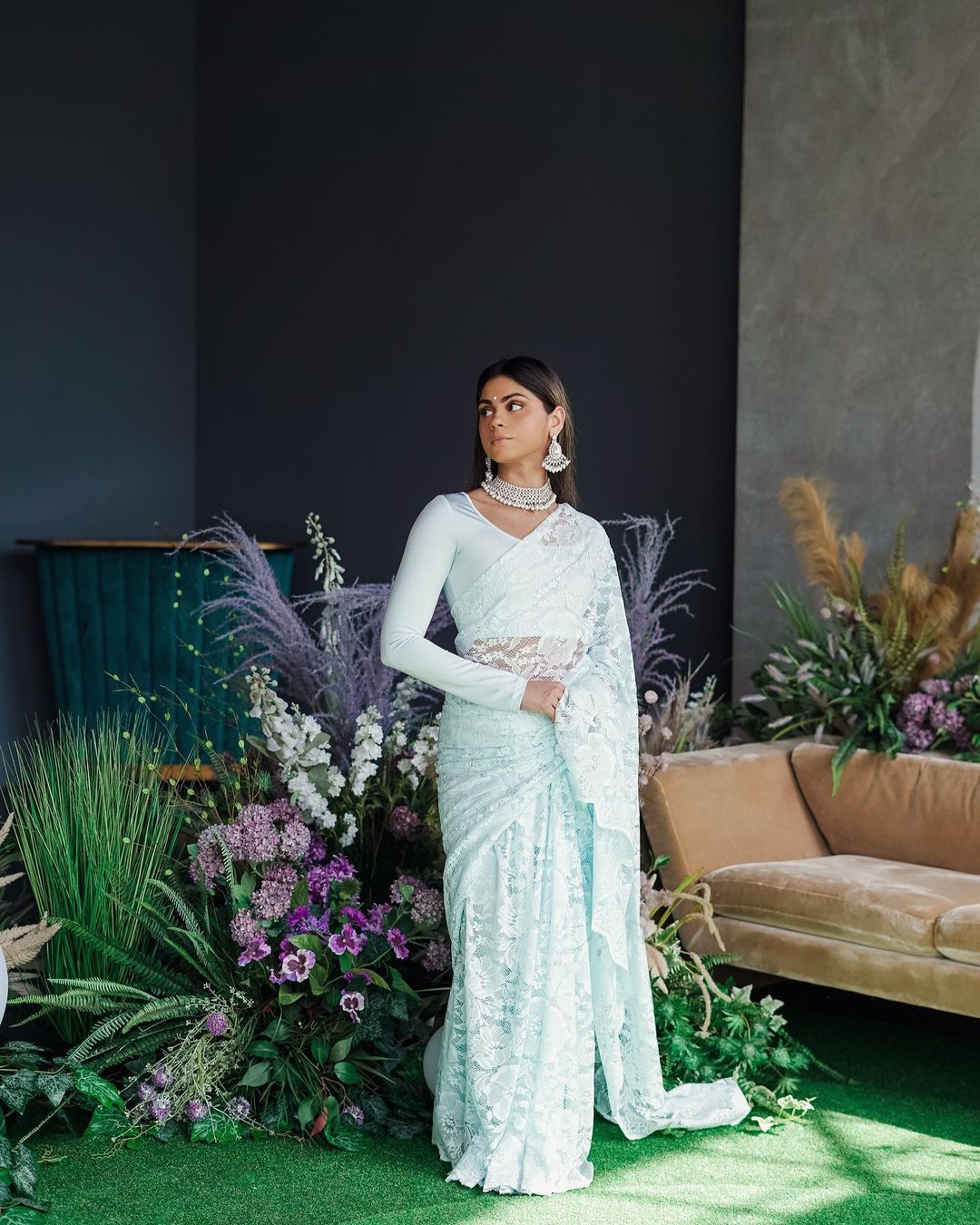 Model draped in a sky blue lace saree with scalloped edging. Model is also wearing a long sleeve crop top and saree petticoat underneath in matching sky blue color. Background is decorated with floral bouquets.