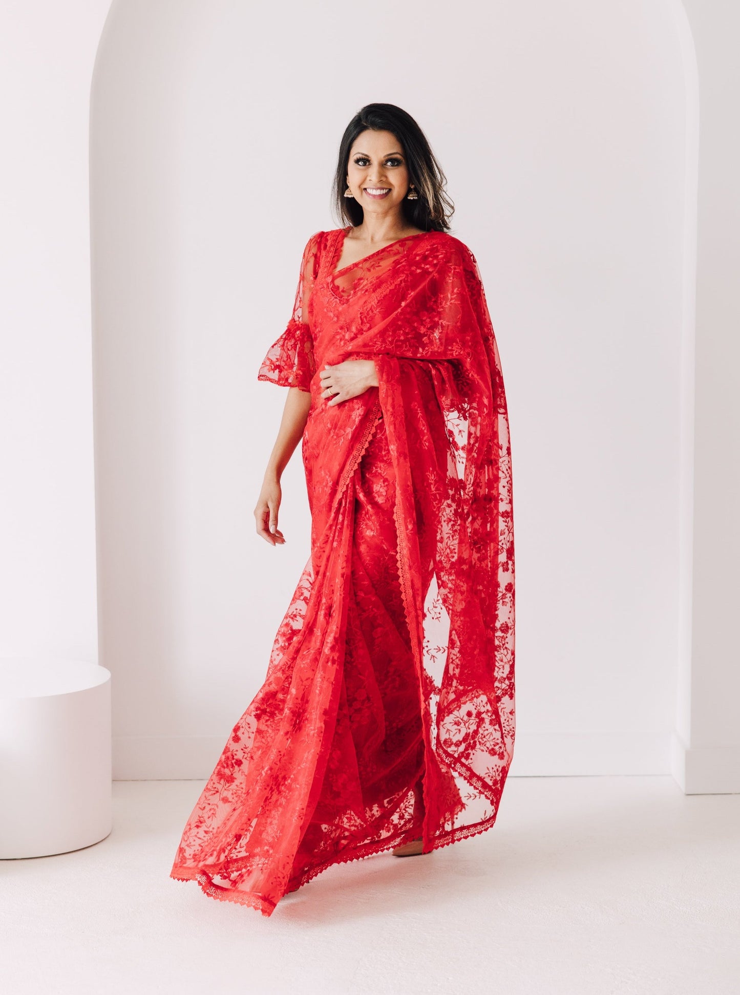 Model draped in a bright red transparent mesh saree with red floral embroidery and a red lace border. Model is wearing a matching red blouse with flared sleeves and a red saree petticoat underneath.