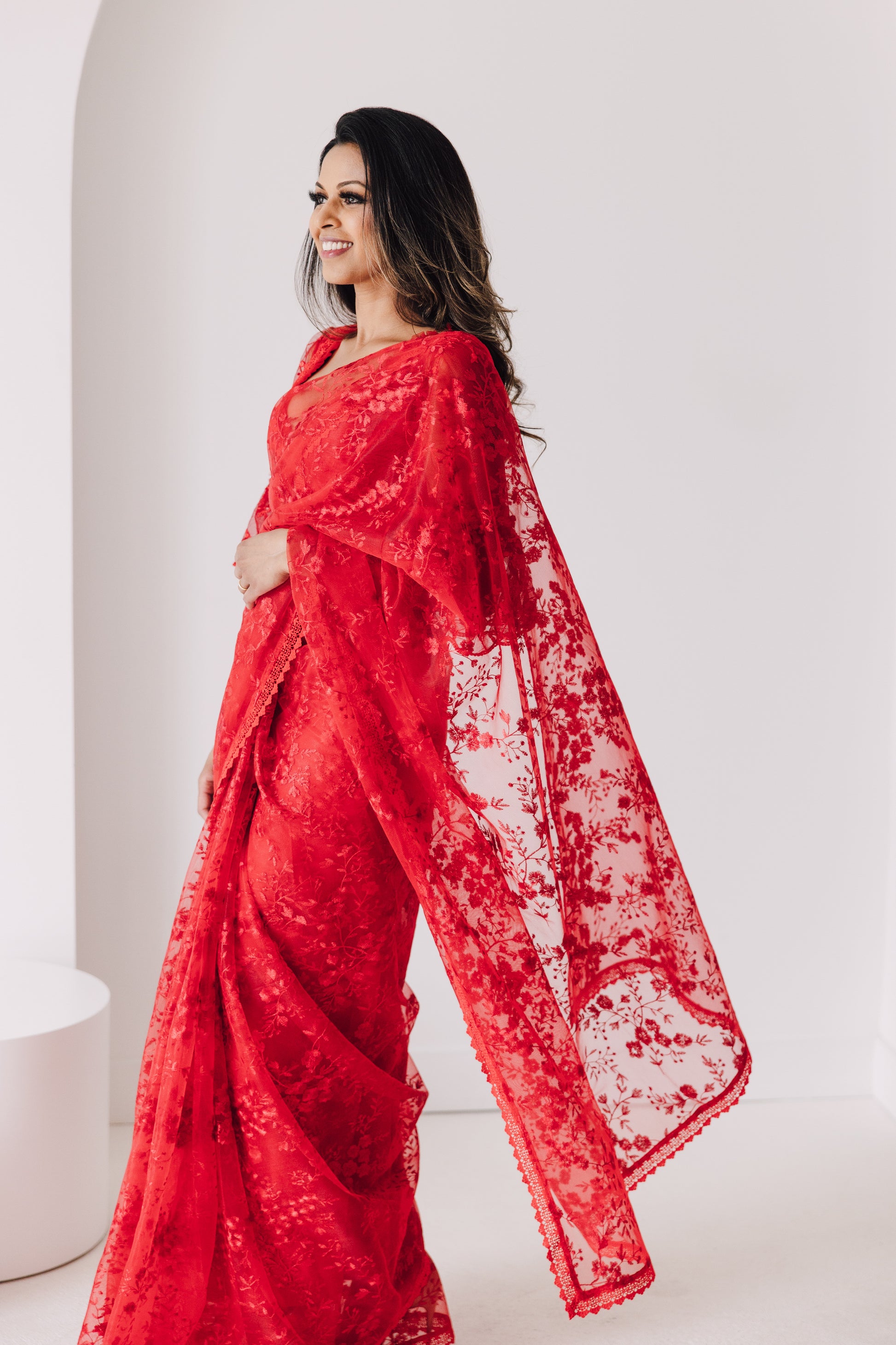 Model draped in a bright red transparent mesh saree with red floral embroidery and a red lace border. Model is wearing a matching red blouse with flared sleeves and a red saree petticoat underneath