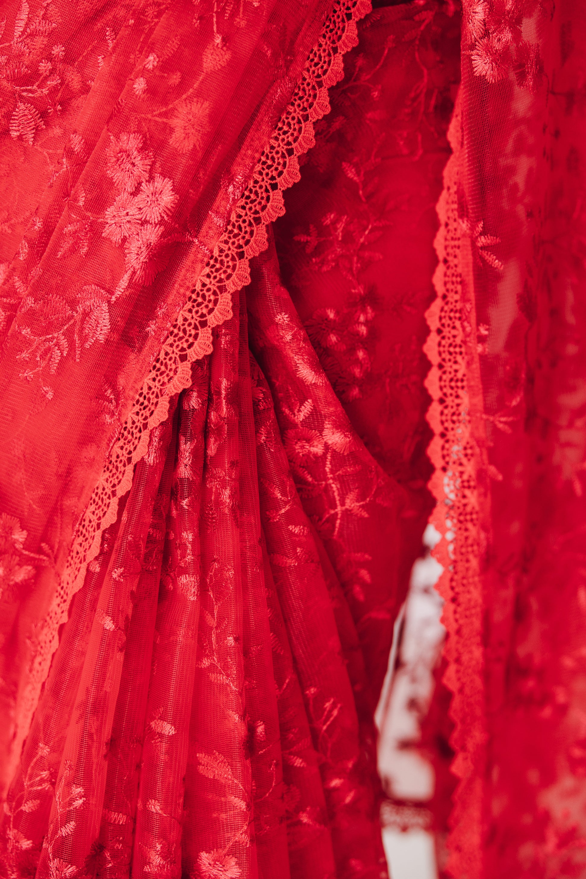 Close up of front pleats on a draped bright red transparent mesh saree with red floral embroidery and a red lace border.