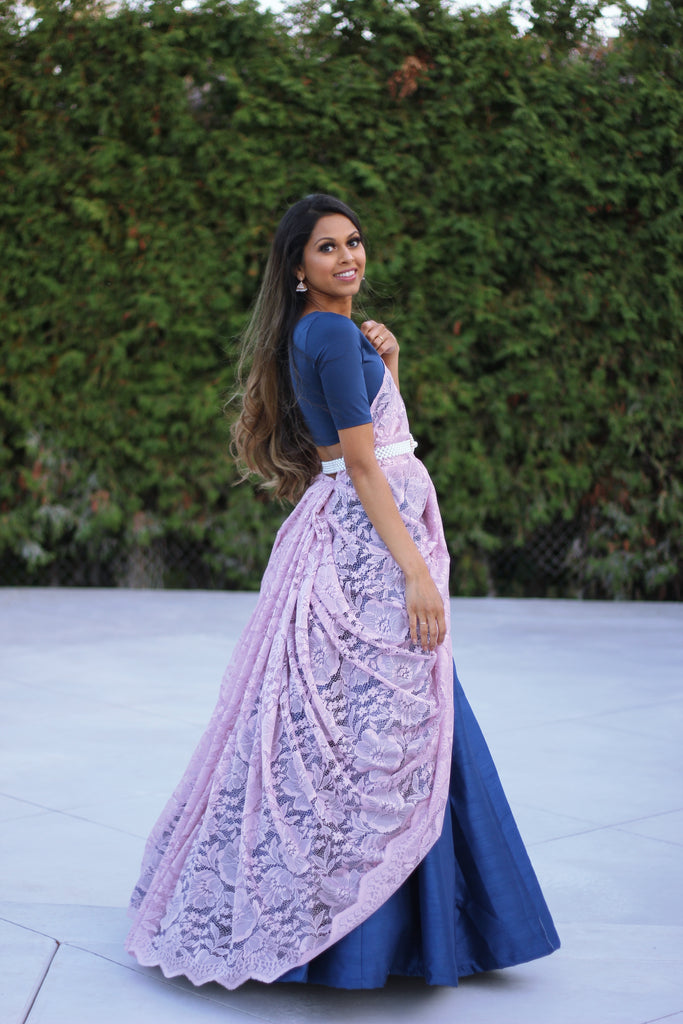 Model draped in a lilac purple lace saree with scalloped edging. The saree is draped over a sapphire blue short sleeve crop top and matching blue cancan skirt. Look is completed by a pearl waist chain.