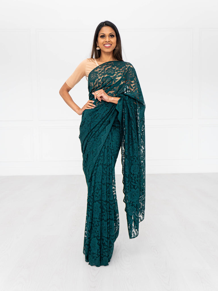 Model draped in an emerald green lace saree with scalloped edging. Model is also wearing a short sleeve champagne colored crop top and saree petticoat underneath in matching emerald green color.
