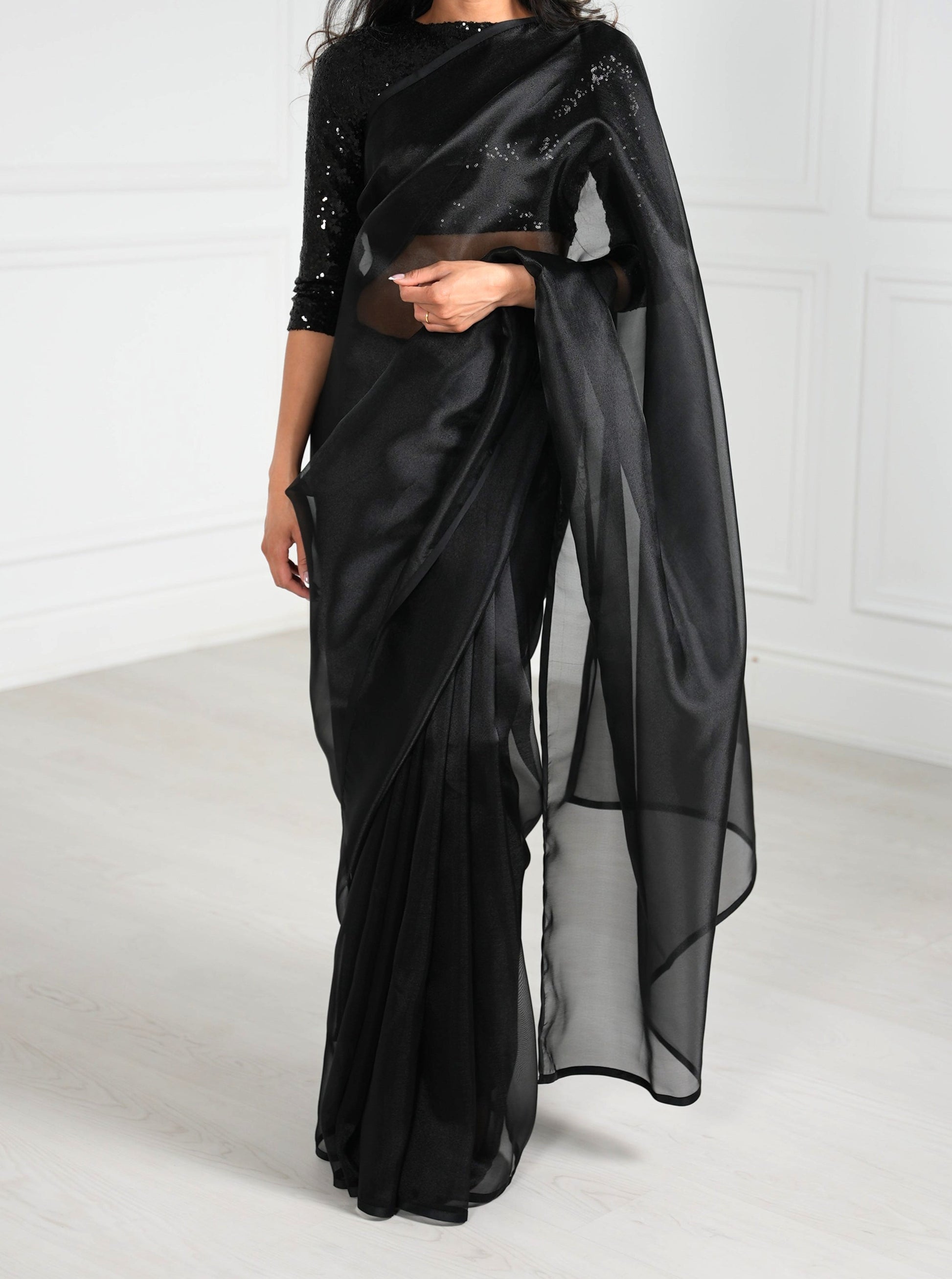 Model draped in a black organza saree with black satin border. Model is also wearing a black 3/4 sleeve sequin crop top.