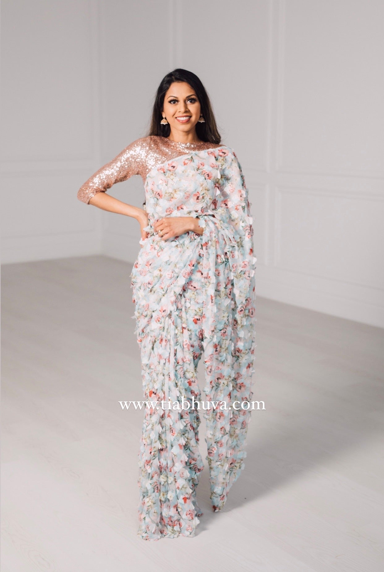 Model draped in a lightweight pale blue saree with a red, green, and pink floral print and 3D petals. Model is also wearing a rose gold high neck 3/4 sleeve sequin crop top.