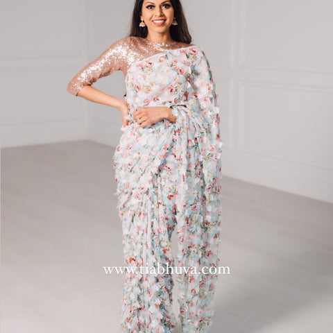 Model draped in a lightweight pale blue saree with a red, green, and pink floral print and 3D petals. Model is also wearing a rose gold high neck 3/4 sleeve sequin crop top.