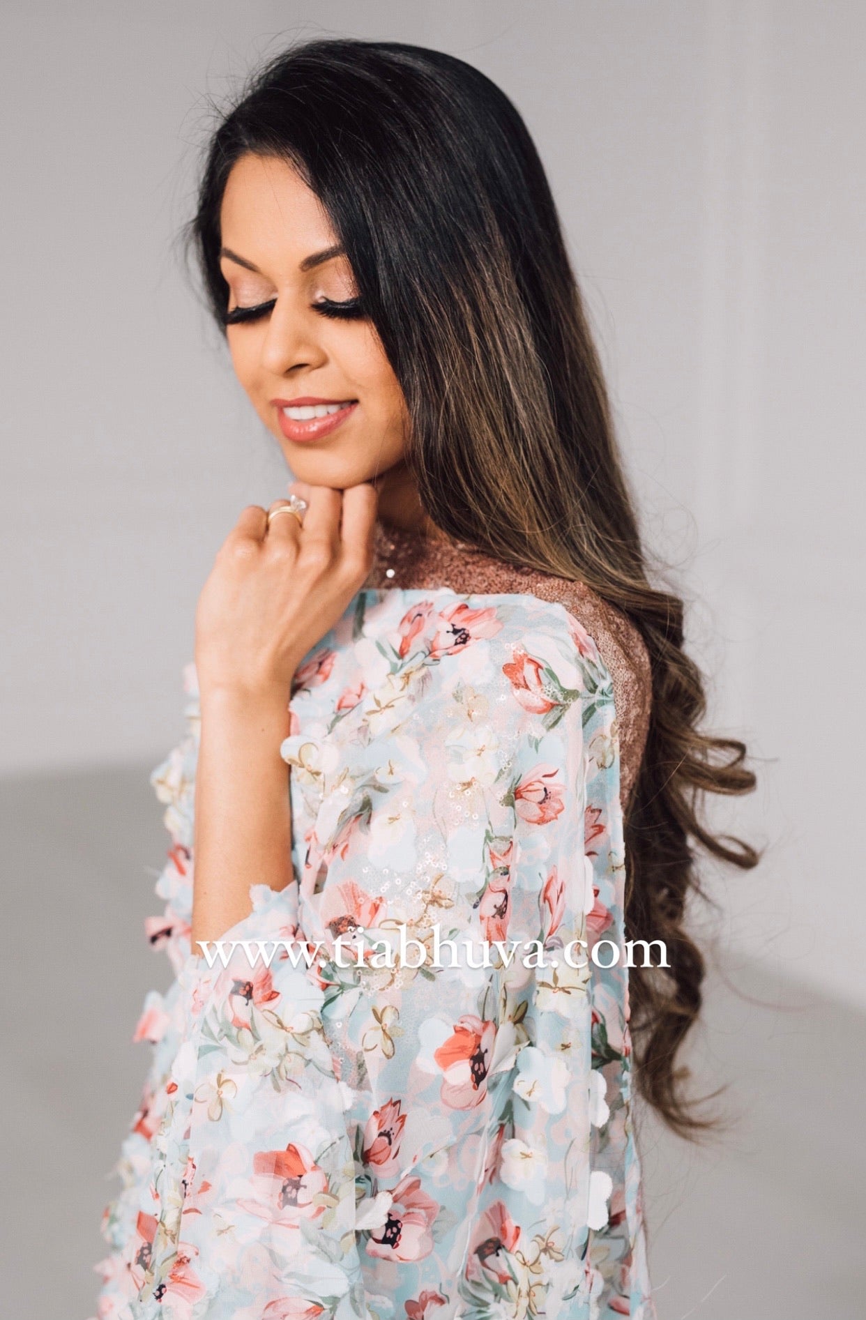 Side view of a model draped in a lightweight pale blue saree with a red, green, and pink floral print and 3D petals. Model is also wearing a rose gold high neck 3/4 sleeve sequin crop top.