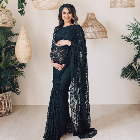 Model holding her baby bump draped in a black lace saree with scalloped edging. Model is also wearing a black long sleeve lace crop top and black saree petticoat underneath.