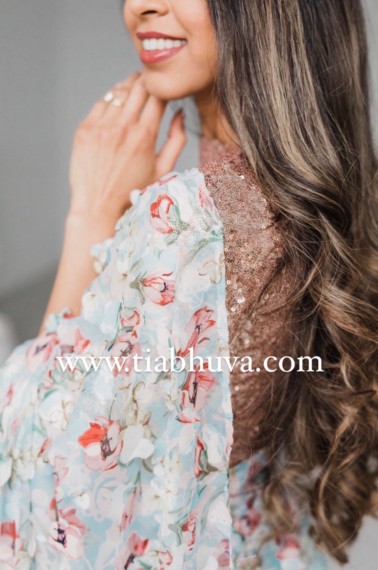 Side view of a model draped in a lightweight pale blue saree with a red, green, and pink floral print and 3D petals. Model is also wearing a rose gold high neck 3/4 sleeve sequin crop top.